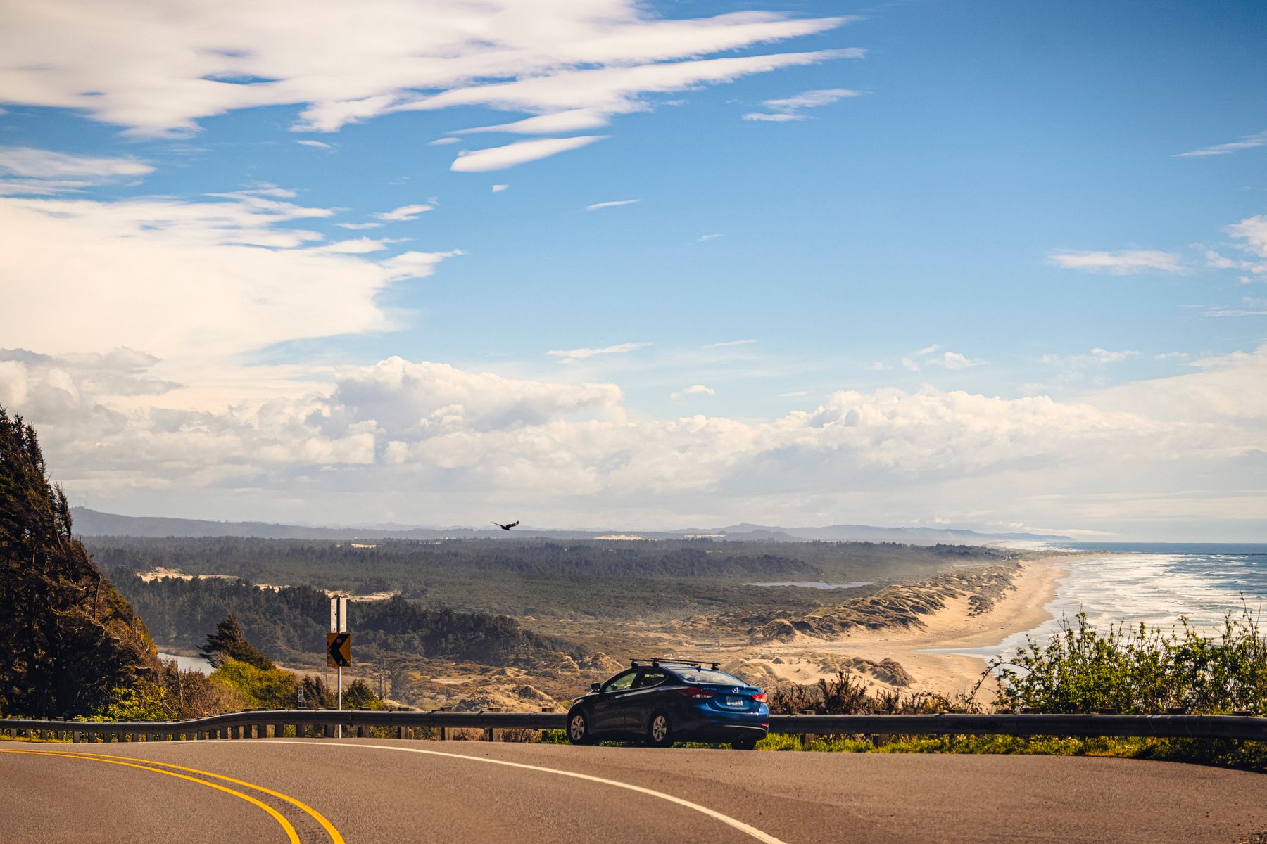 Blue car parked on an Oregon coastal highway above sand dunes and the Pacific